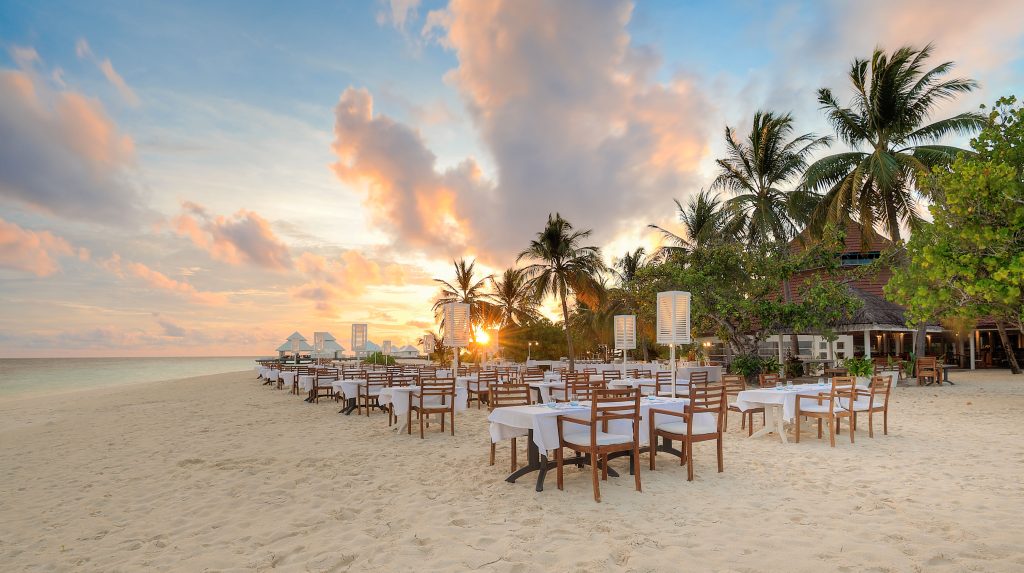 Outdoor restaurant setup on a sandy beach at sunset, with wooden tables and chairs covered in white cloths, surrounded by palm trees, and the ocean in the background under a partly cloudy sky.