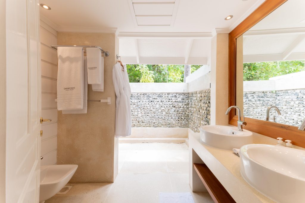 Bright, modern bathroom with two white sinks, large mirror, neatly folded towels, and an outdoor shower area surrounded by stone walls and greenery. A white robe hangs by the open shower space.