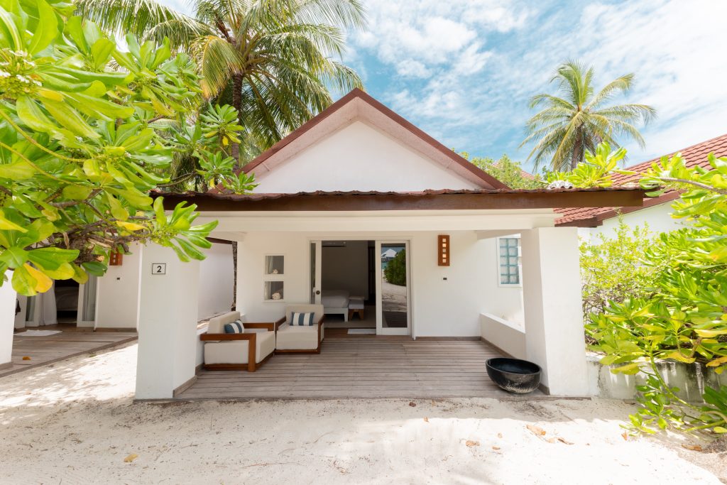 White beach villa with a shaded patio, outdoor seating, and tropical plants around. The villa has large glass doors leading to a bright interior, with palm trees and a blue sky in the background.