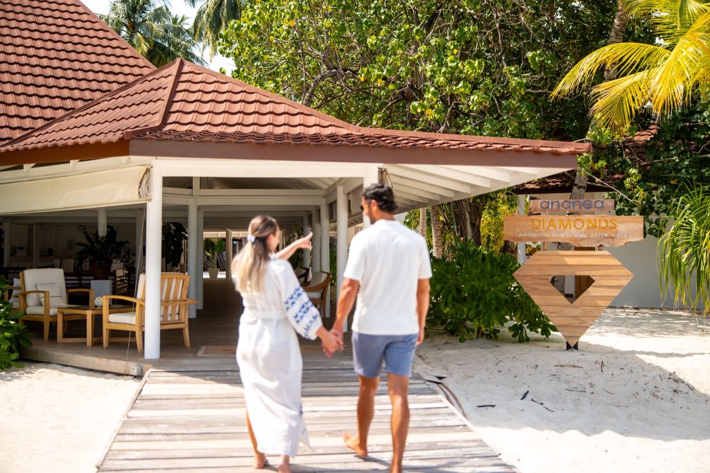 A couple holding hands walks toward a tropical beach resort with a red-tiled roof and a Diamonds sign surrounded by lush green trees and white sand.