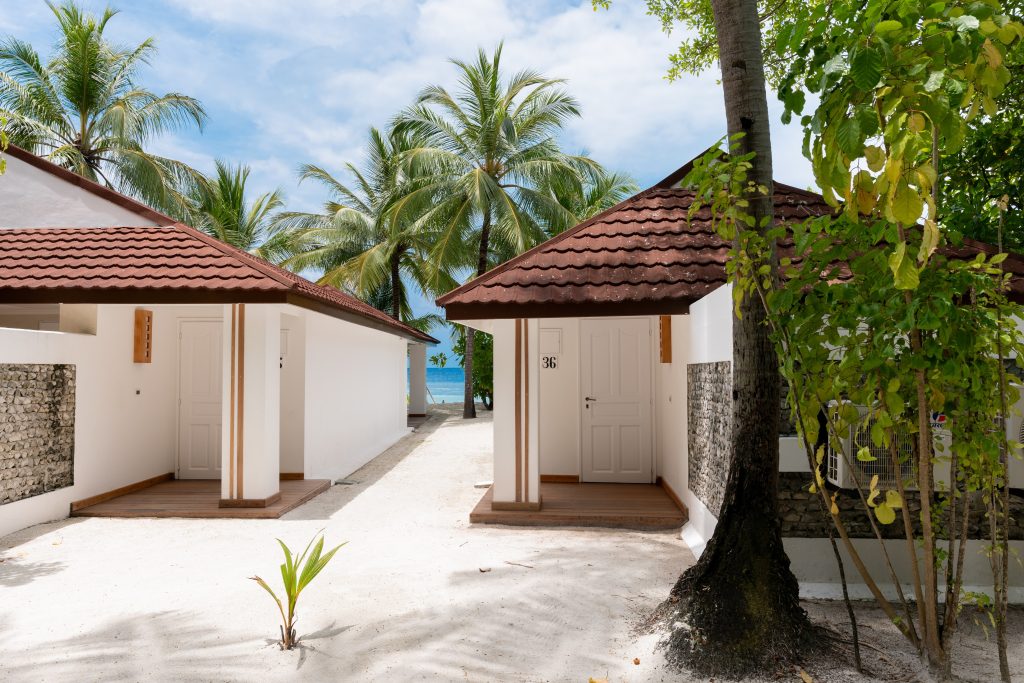 Two small white villas with red tile roofs stand side by side on a sandy path lined with palm trees, with the ocean and blue sky visible in the background.