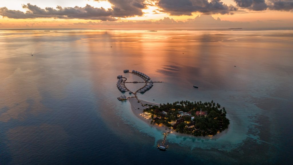 Aerial view of a small tropical island resort at sunset, surrounded by calm ocean waters, overwater villas, and palm trees, with warm lights illuminating pathways on the island.