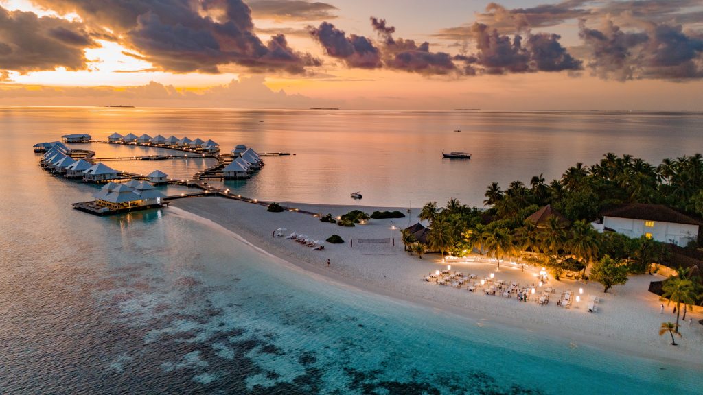 Aerial view of a tropical island resort at sunset, featuring overwater villas, white sandy beaches, turquoise water, palm trees, and outdoor dining areas illuminated by warm lights.