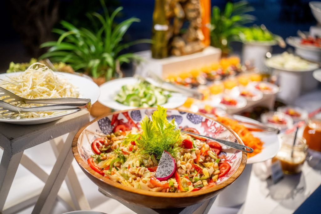 A colorful buffet table features a large salad with tomatoes, lettuce, and dragon fruit in the foreground, surrounded by various fresh vegetables, garnishes, and tongs, with plants and appetizers in the background.