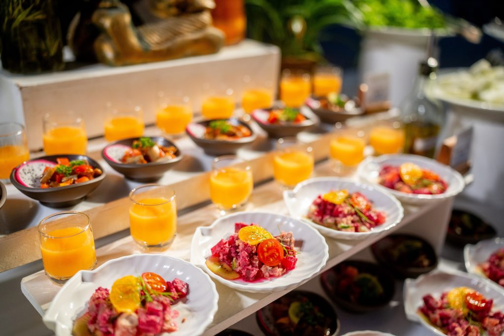 A display of small plates with colorful salads, topped with tomatoes and greens, arranged on white tiers. Glasses of orange juice and bowls of mixed appetizers are also neatly placed in rows.