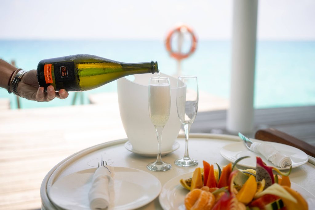 A hand pours champagne into a glass beside a white ice bucket on a table set with plates, napkins, and a colorful fruit platter, overlooking a bright blue ocean with a life preserver in the background.