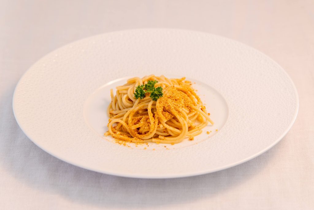 A white plate with a serving of spaghetti topped with grated yellow cheese and garnished with a small sprig of parsley, set on a light-colored tablecloth.
