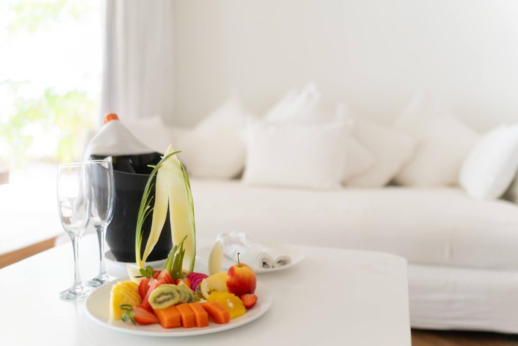 A plate of assorted fresh fruit and desserts sits on a white table next to two empty wine glasses and a wine bottle in an ice bucket, with a white sofa and pillows in the background.