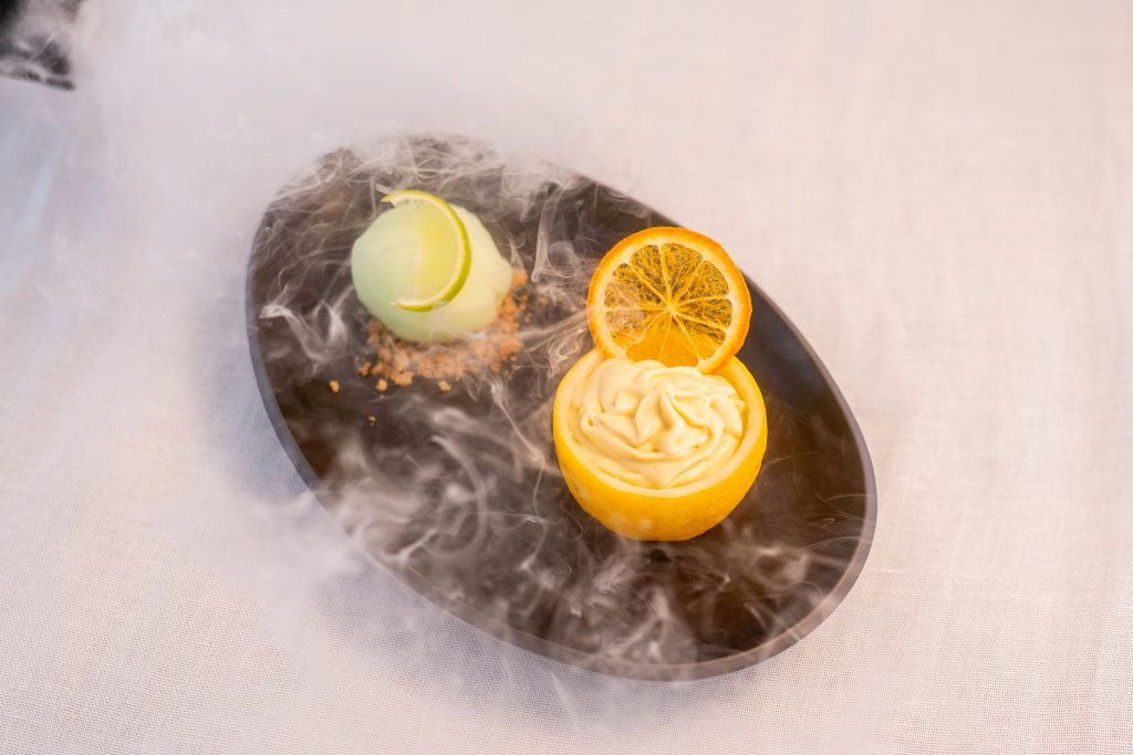 A black oval plate with a lemon half filled with cream and a dried lemon slice, next to a green dessert ball garnished with a lime slice and crushed nuts, surrounded by wisps of white smoke.