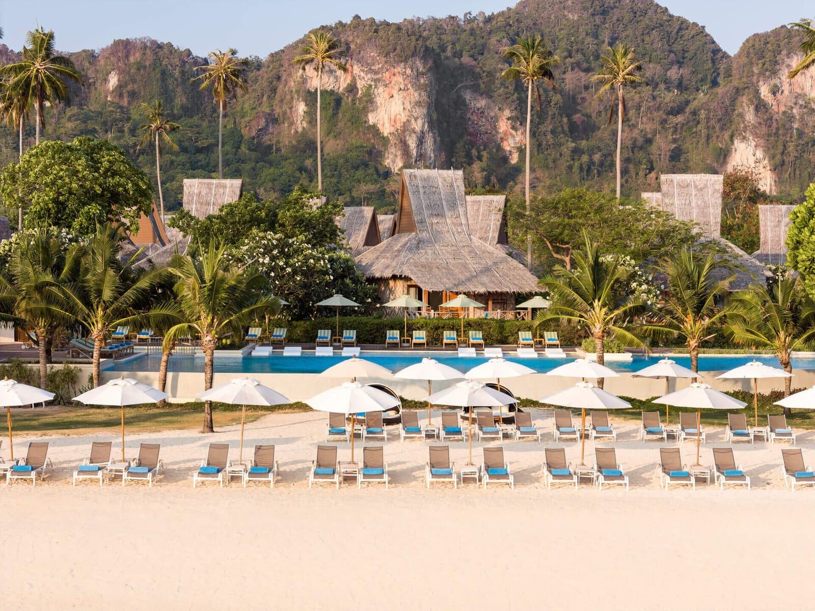 Rows of lounge chairs and white umbrellas line a sandy beach facing a resort pool, with tropical trees and thatched-roof buildings, set against a backdrop of lush green hills and rocky cliffs.