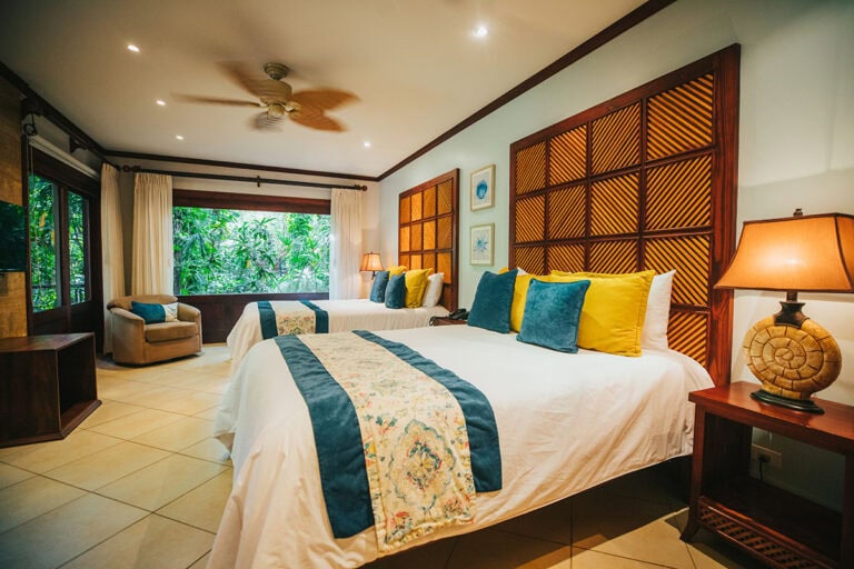 A bright hotel room with two double beds, colorful pillows, warm lighting, a large window with a forest view, a wooden armchair, and tiled floors.