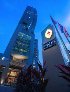 Night view of the Eastin Grand Hotel, featuring a tall modern building, illuminated signage, and three flags on poles. Decorative plants are in the foreground against a blue evening sky.