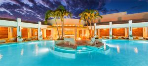 A luxurious, illuminated pool with palm trees and lounge seating in the center, surrounded by modern cabanas and warm lighting under a dramatic evening sky.