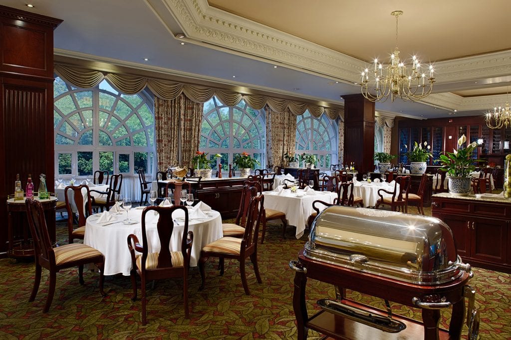 Elegant restaurant dining room with large arched windows, round tables covered with white tablecloths, classic wooden chairs, chandeliers, and buffet serving station in the foreground.