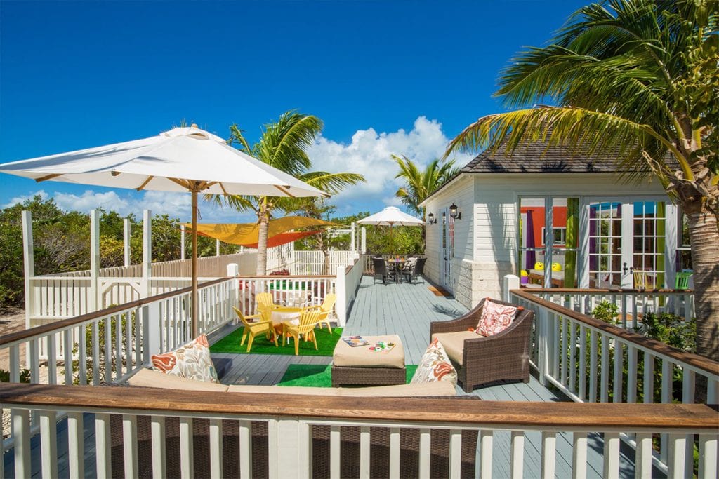 A sunny outdoor deck with wicker sofas, colorful cushions, and tables, surrounded by white railings. Palm trees and umbrellas provide shade, and a beach house is visible under a bright blue sky with scattered clouds.