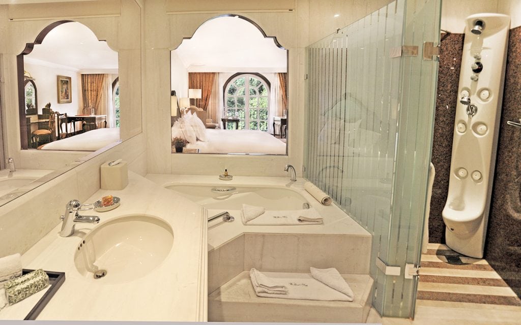 Elegant hotel bathroom with a large mirror, sink, corner bathtub, modern shower panel, and towels. An arched window shows a view into a cozy bedroom with a large bed and bright natural lighting.