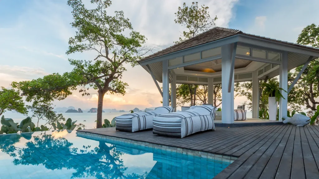 A luxurious poolside cabana with striped lounge chairs overlooks a scenic view of the ocean and distant islands, surrounded by trees at sunset.