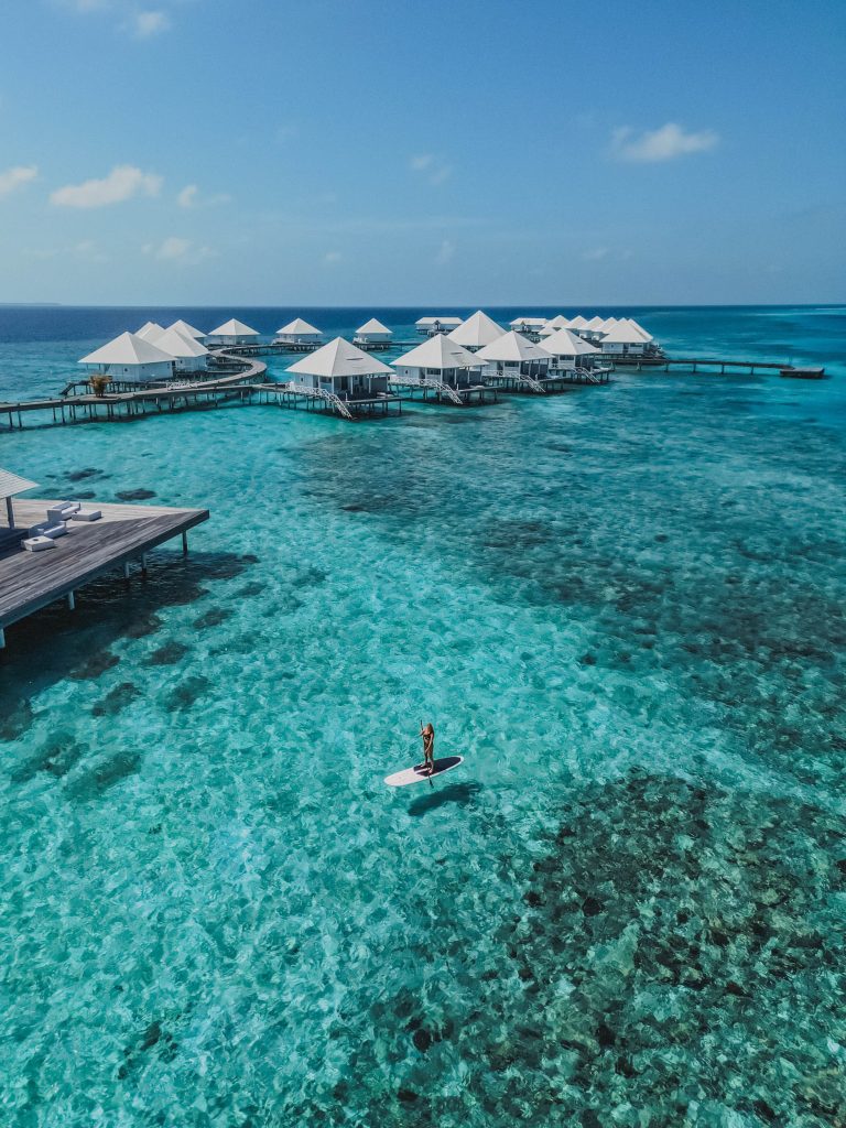 A person paddleboards on clear turquoise water near white-roofed overwater villas, surrounded by coral reefs under a bright blue sky.