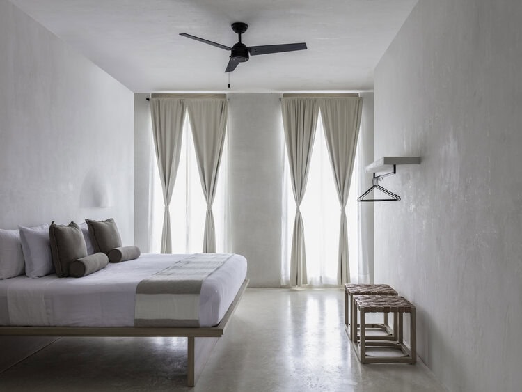 Minimalist bedroom with white walls, a double bed with gray and white bedding, two tall windows with sheer curtains, a ceiling fan, a wall-mounted rack with hangers, and two woven stools.