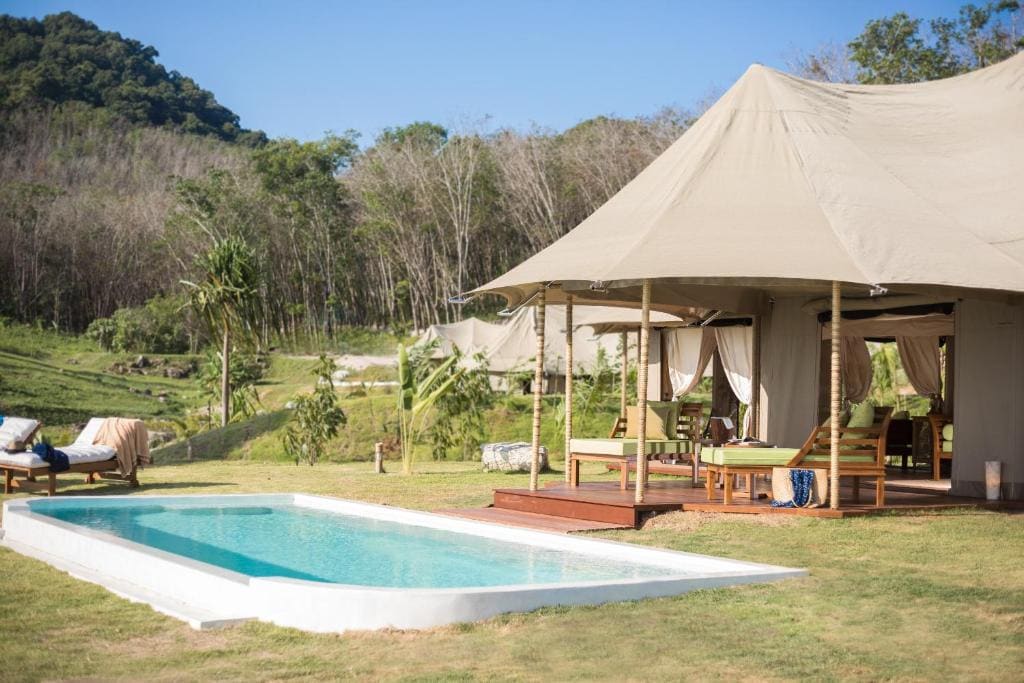 A luxury tent with a shaded porch and lounge chairs sits beside a small rectangular pool on grassy grounds, surrounded by lush trees and hills under a clear blue sky.