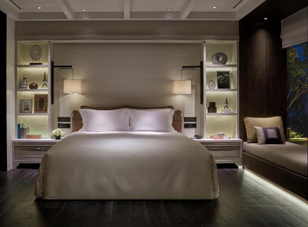 A modern, cozy bedroom with a large bed centered against a paneled wall, flanked by shelves displaying decor. Two wall lamps provide soft lighting, and a window seat with cushions sits to the right.