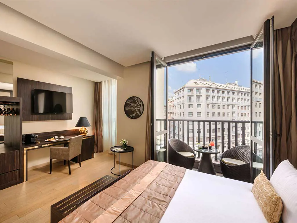 Modern hotel room with a large bed, desk, TV, and balcony. The balcony has two chairs and a table, overlooking city buildings under a blue sky. The room features warm, neutral decor and natural light from large windows.