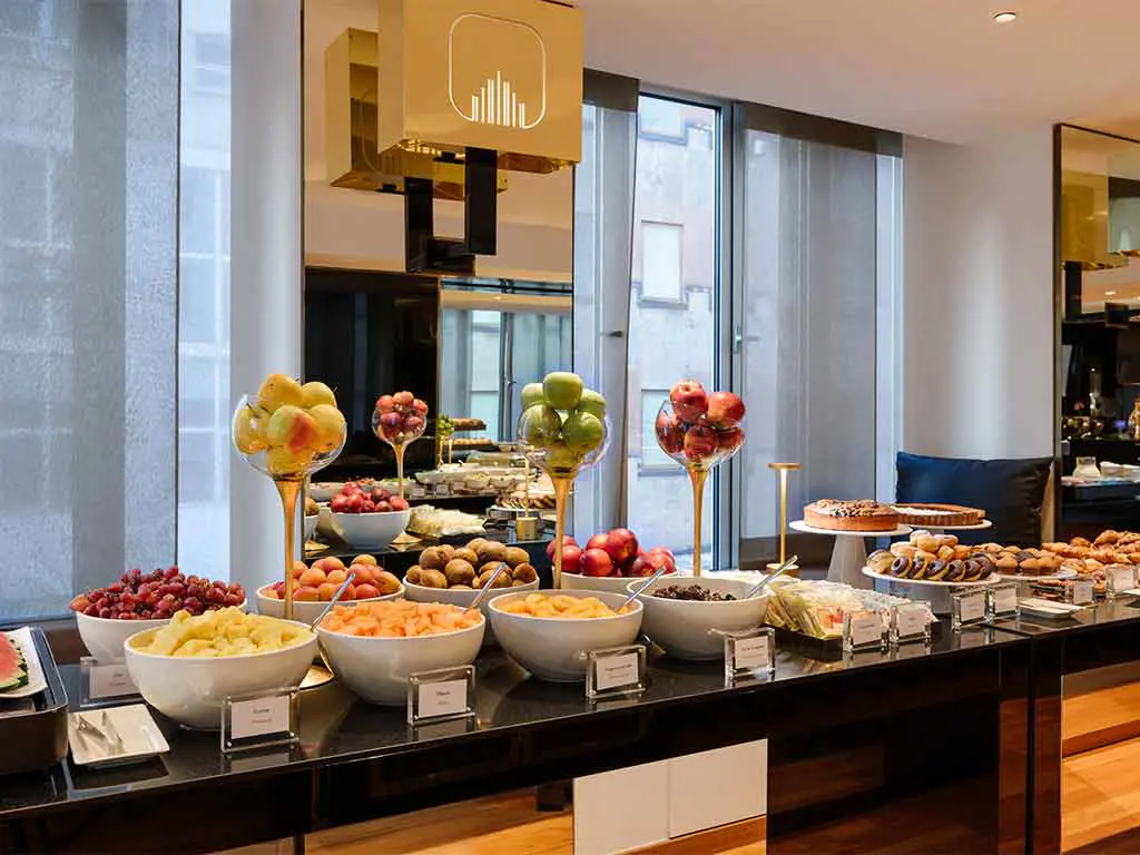 A buffet table features bowls of fresh fruits, assorted pastries, and desserts, displayed in a modern, well-lit dining area with large windows and mirrors.