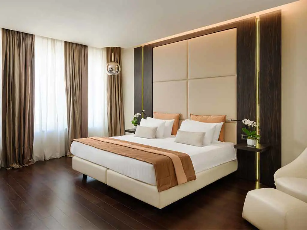 Modern hotel room with a large bed, white and beige bedding, two bedside tables with white flowers, brown curtains, wooden floor, and neutral-toned walls with soft lighting.