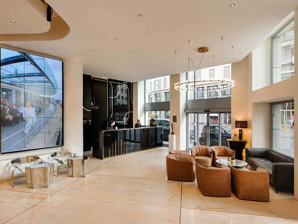 Modern hotel lobby with a black reception desk, two brown leather chairs, a sofa, a circular chandelier, wall art, and large windows letting in natural light. Two people stand at the reception counter.