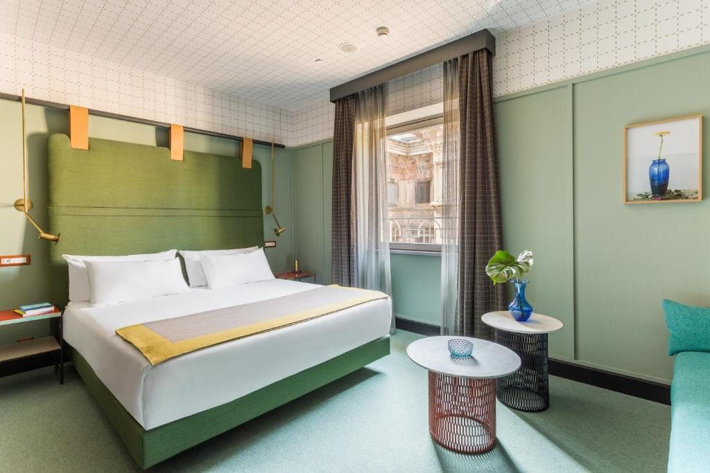 Modern hotel room with a large bed, green padded headboard, two round side tables, a potted plant, a framed artwork, and a large window with sheer curtains letting in natural light.