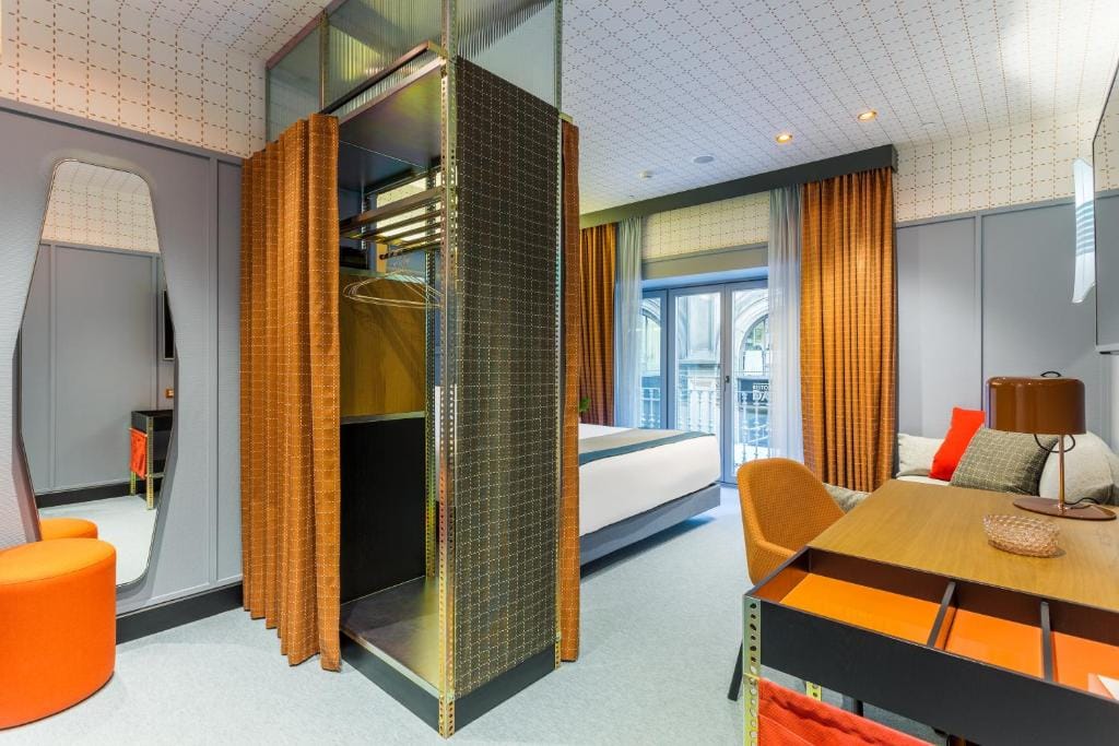Modern hotel room with orange curtains, a double bed, a desk with an orange chair, an open wardrobe, a wall mirror, and a seating area with an orange ottoman and a sofa. Large windows let in natural light.