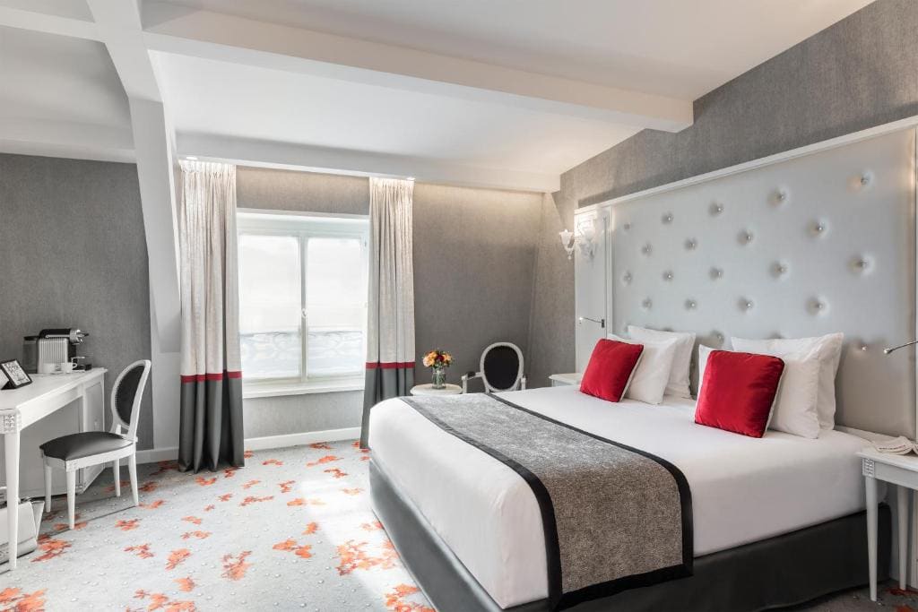 A modern hotel room with a large bed, gray and white decor, red accent pillows, a tufted headboard, a desk with a chair, a small round table with flowers, and a window with curtains letting in natural light.