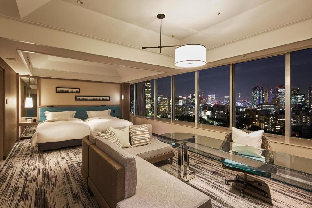 A modern hotel room with two beds, a sofa, a desk, and large windows showcasing a city skyline at night with illuminated buildings. The room is stylishly decorated in neutral tones.