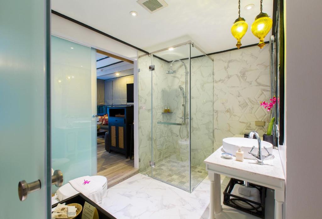 Modern bathroom with a glass shower, marble-style walls and floor, a white sink on a vanity, hanging yellow pendant lights, and pink flowers near the mirror. Part of a bedroom is visible through a frosted sliding door.