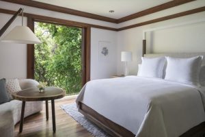 A bright bedroom with a large bed, white linens, a wooden floor, a side table with a lamp, a sofa, a round wooden coffee table, artwork on the wall, and a balcony door open to lush greenery outside.