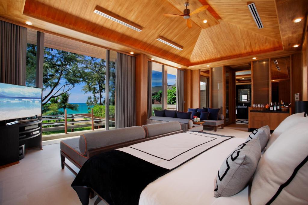 Spacious, modern bedroom with wood-paneled ceiling, large windows overlooking the ocean and greenery, a cozy bed, sofa, flat-screen TV, and elegant decor. Natural light fills the stylish, relaxing space.