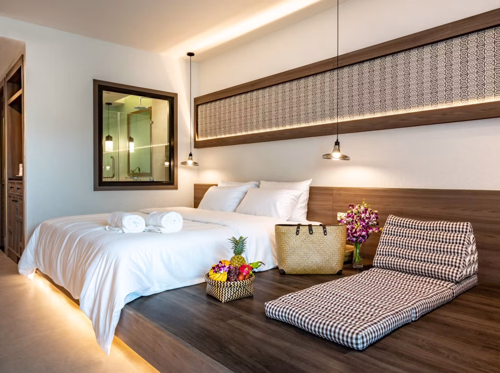 A modern hotel room with a neatly made double bed, white linens, rolled towels, a basket of fruit, a woven bag, a checkered floor cushion, and a bouquet of purple flowers on a wooden platform.