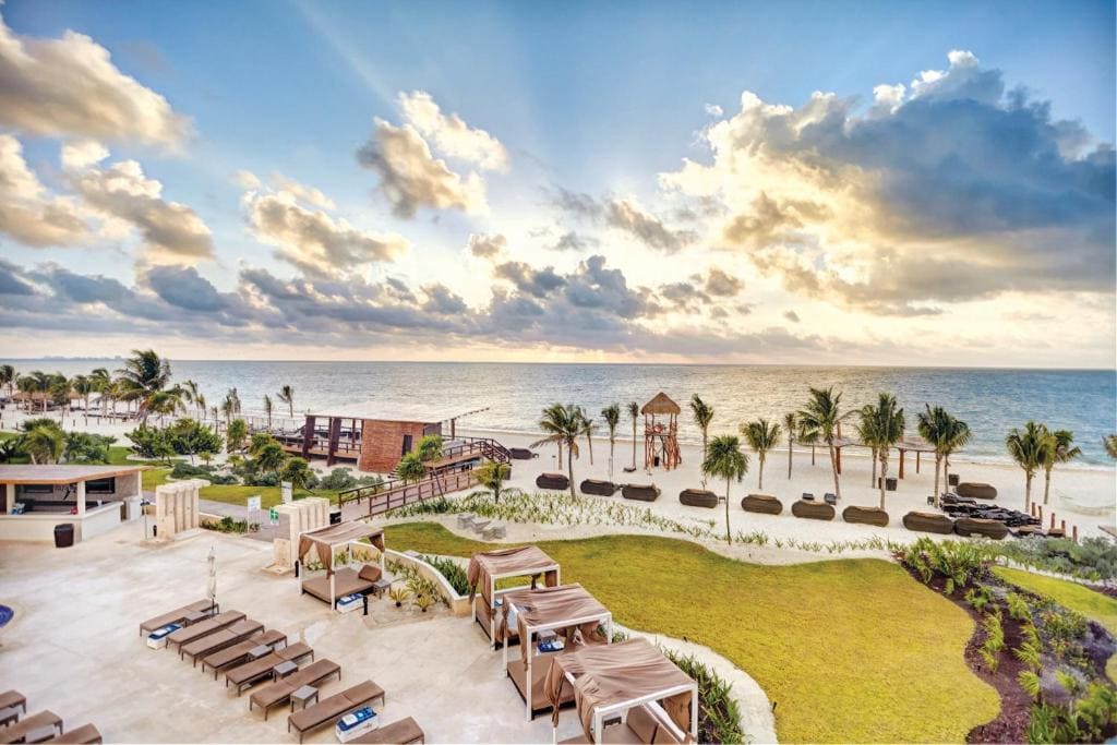 A view of a tropical beach resort with lounge chairs, cabanas, palm trees, and a sandy shoreline beside the ocean under a partly cloudy, sunlit sky.