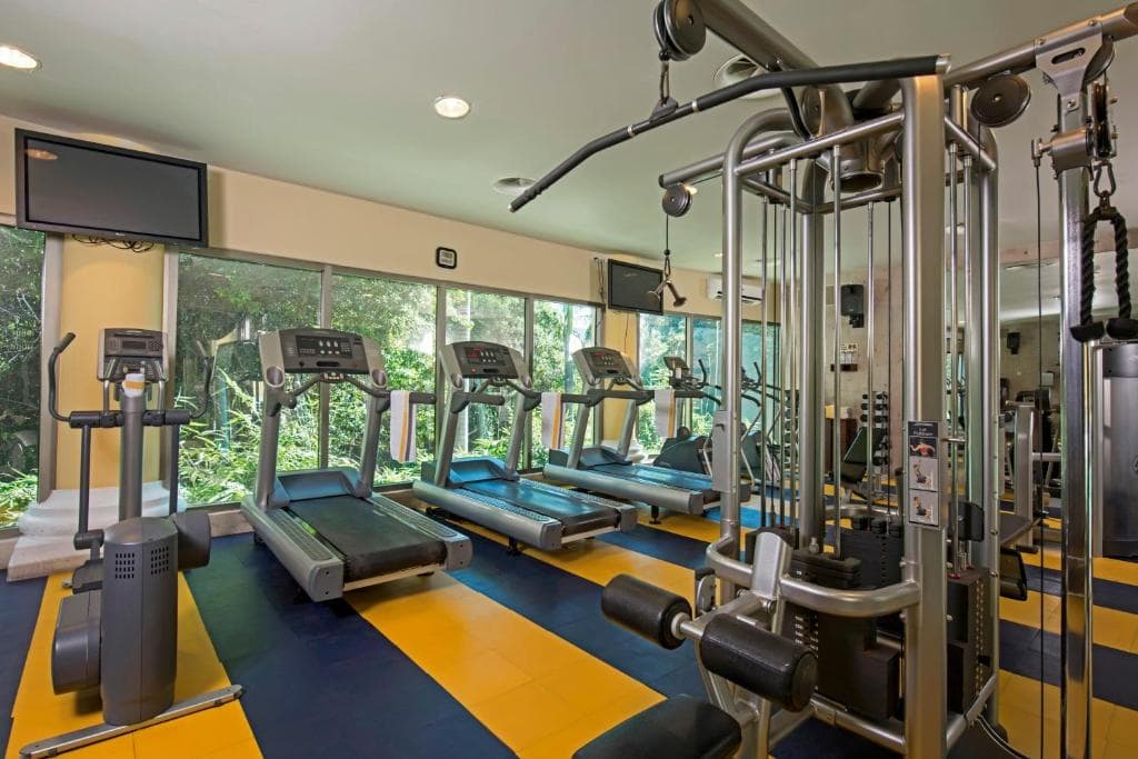 A modern gym with treadmills, exercise bikes, and weight machines on a blue and yellow striped floor. Large windows reveal greenery outside, and TVs are mounted on the walls. The space is well-lit and organized.