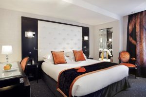Modern hotel room with a large white tufted headboard, a double bed with white linens, orange and black accents, a desk with a lamp, a mirror, and decorative vases; large curtains and soft lighting.