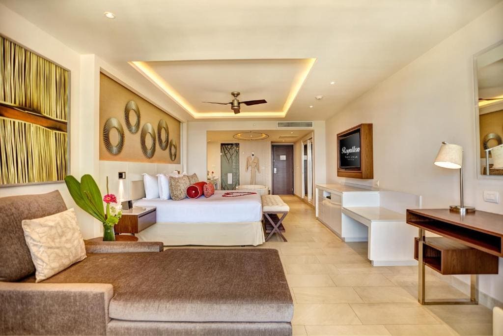 Spacious, modern hotel room with a large bed, chaise lounge, desk, wall-mounted TV, and stylish decor. Warm lighting and neutral tones create a relaxing atmosphere. A plant and decorative art add a touch of elegance.