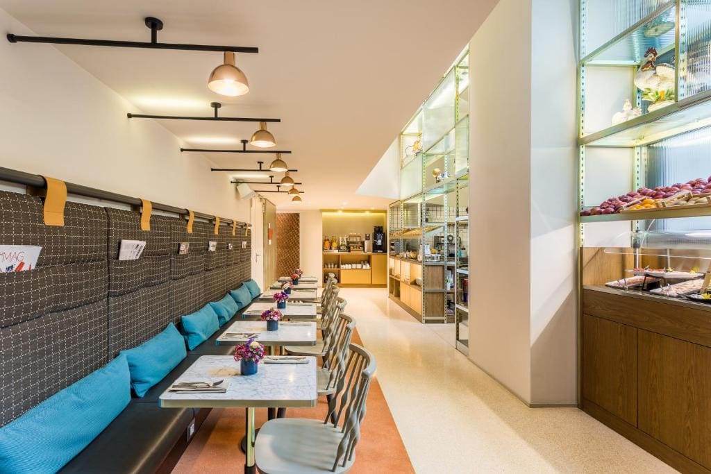 Modern café interior with tables and striped chairs on the left, blue cushions along a bench, small flower vases on tables, and a brightly lit display with pastries and fruit on the right. Warm lighting creates a cozy atmosphere.