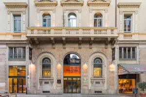 Facade of a grand, historic building with arched windows and ornate architectural details. The entrance reads “Room Mate Hotel,” flanked by large glass doors, with a restaurant to the right and a balcony above.