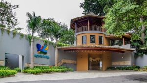 The entrance of a modern lodge with a circular tower, wooden balcony, and large gate. The sign reads Lágrima Lodge. Lush greenery and palm trees surround the building.