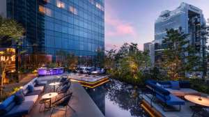 A modern rooftop lounge at sunset features blue sofas, round tables, and glowing lights, surrounded by greenery and reflected in a shallow pool, with tall glass buildings in the background.