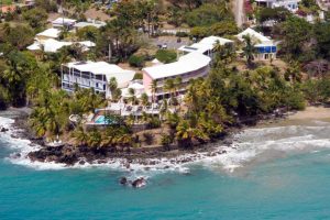 Aerial view of a coastal resort with white and pink buildings surrounded by palm trees, located on a rocky shoreline next to turquoise ocean water and a small sandy beach.