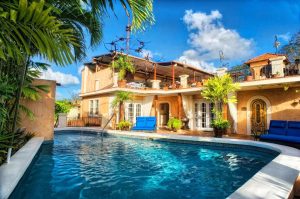 A tropical villa with tan walls, balconies, and blue sofas surrounds a sparkling swimming pool, framed by palm trees and lush plants, under a bright blue sky with scattered clouds.
