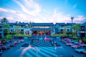 A luxurious resort at sunset features a central building, surrounding villas, a pool with a pink flamingo float, and lounge chairs with pink cushions, all set among palm trees and decorative lights.
