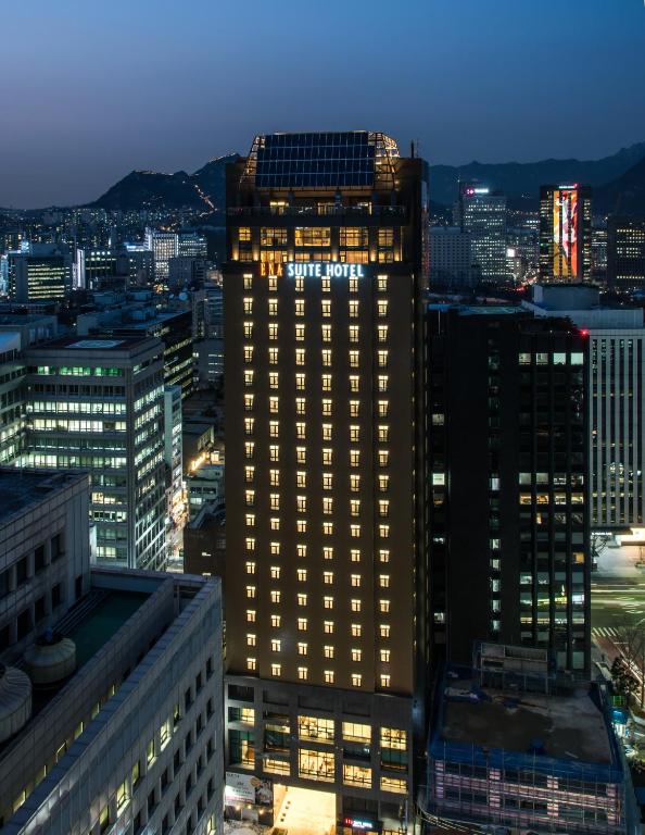 A tall hotel building named Suite Hotel is illuminated at night amidst city skyscrapers, with distant mountains visible under a dusky sky.
