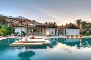 Luxury tropical villa with a thatched roof, large swimming pool, sun loungers, pool umbrellas, and lush palm trees at sunset. The outdoor bar and seating area create a relaxing resort atmosphere.